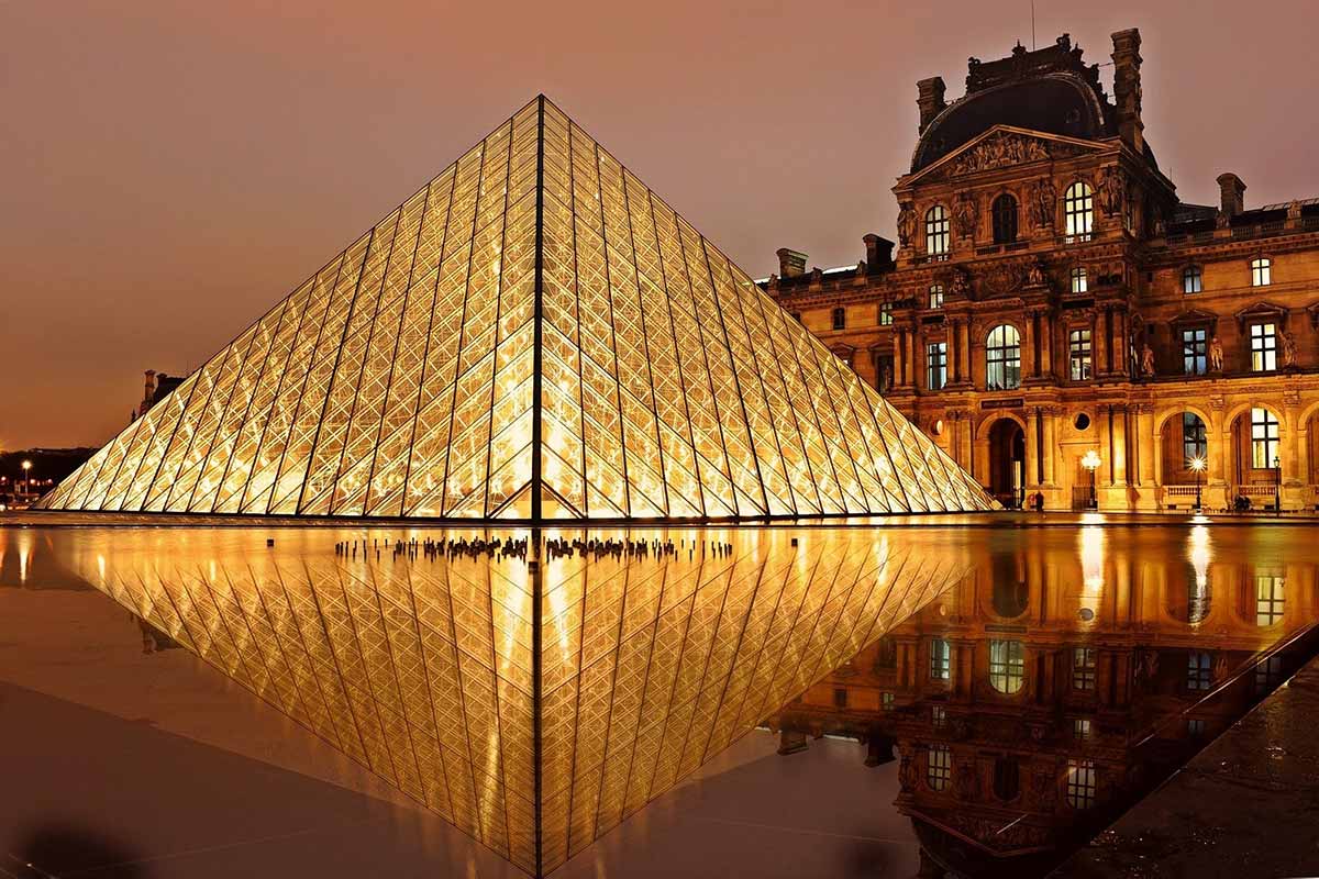 Illuminated Louvre Pyramid at dusk with reflection on water, showcasing the juxtaposition of modern and historical architecture in Paris, France