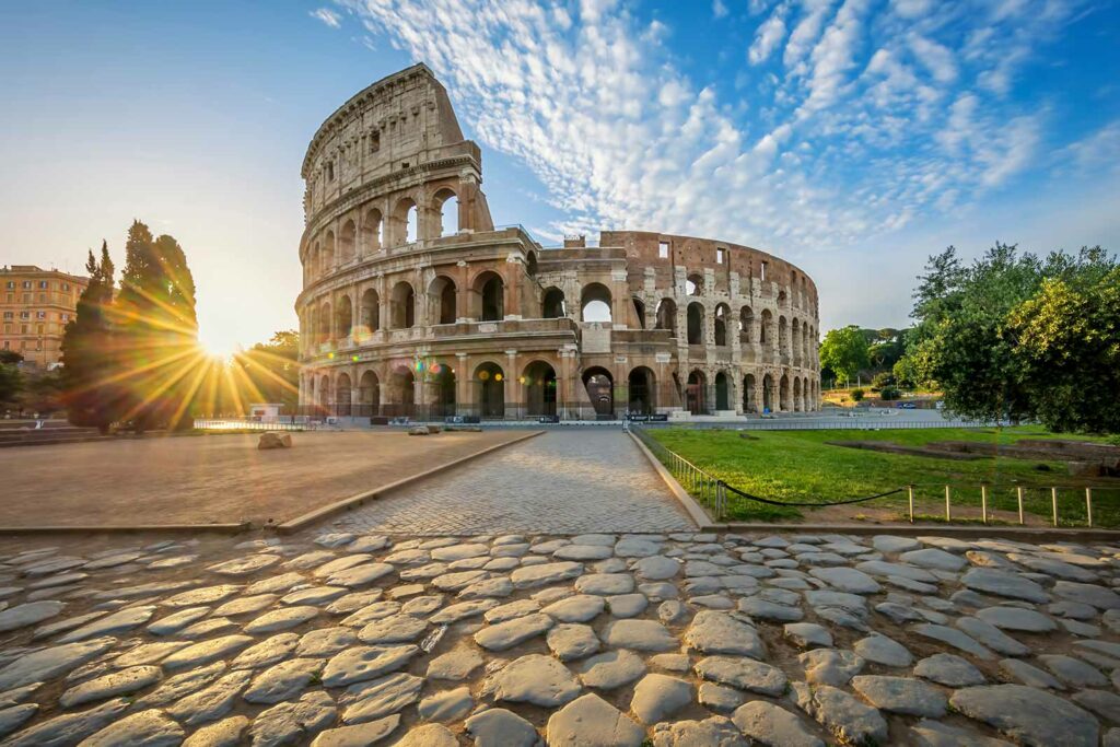 Sunrise over the historic Colosseum in Rome, Italy, showcasing the ancient amphitheater's arches and stonework against a blue sky with scattered clouds.