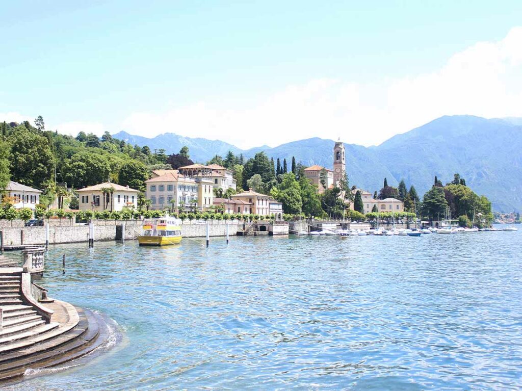 Serene view of Lake Como, Italy showcasing sunlit villas, a charming stone bell tower, and a tour boat near a stepped lakeside promenade