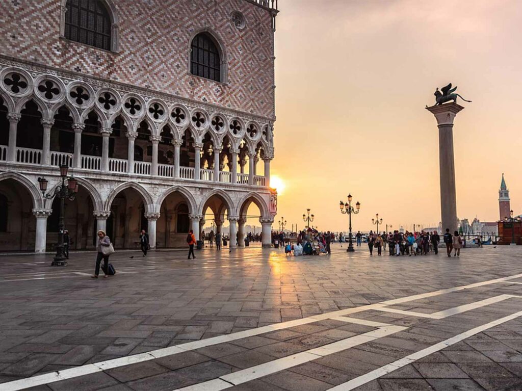 Sunset view of Doge's Palace in Venice with tourists and historic architecture, highlighting the iconic facade and column with a statue.
