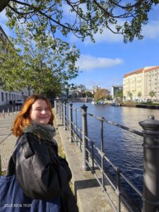 Katie from Luxury France Tours smiling by a scenic river promenade in Berlin with historical buildings and a tree-lined walkway in the background