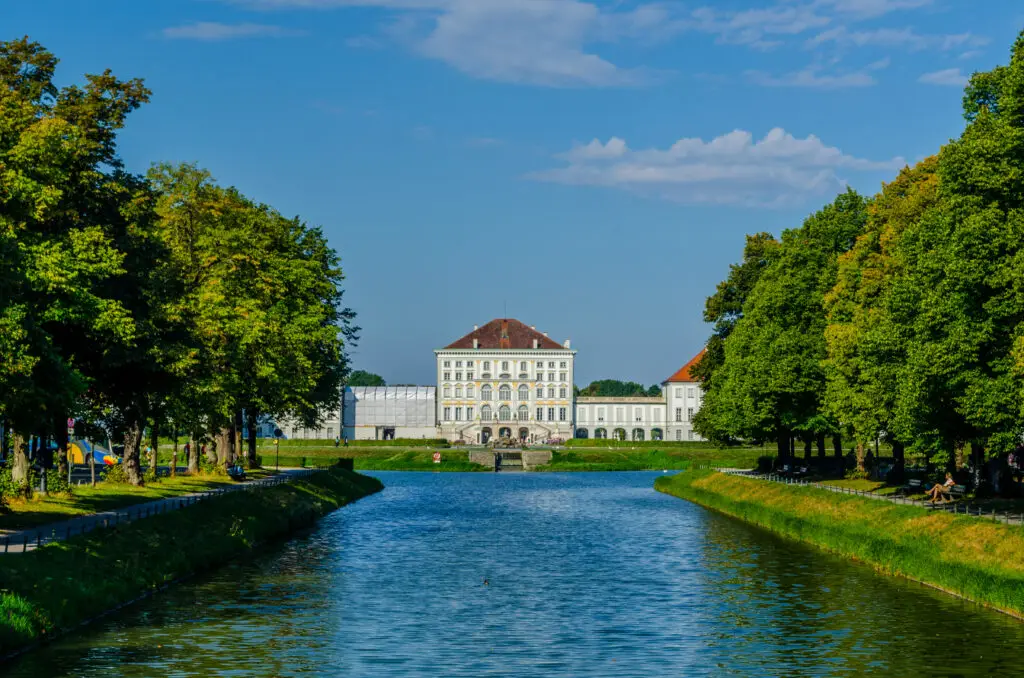 Scenic view of the Nymphenburg Palace in Munich with a frontal facade reflected in the calm canal waters, surrounded by lush green trees under a clear blue sky.