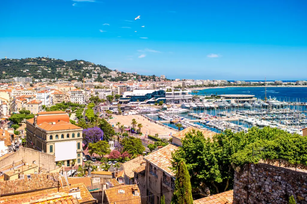 Panoramic view of Cannes city on the French Riviera, showcasing the busy yacht-filled marina, vibrant cityscape, and lush green hills under a clear blue sky.
