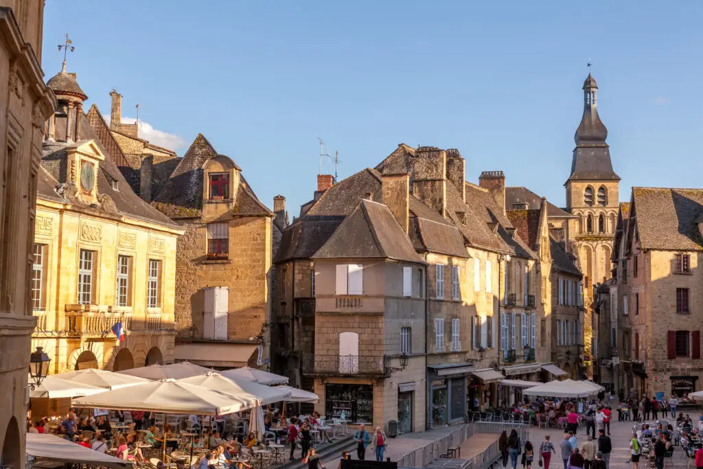Sunny day in Sarlat-la-Canéda, showcasing bustling medieval market square with historic stone buildings and lively cafes filled with tourists in Dordogne, France.