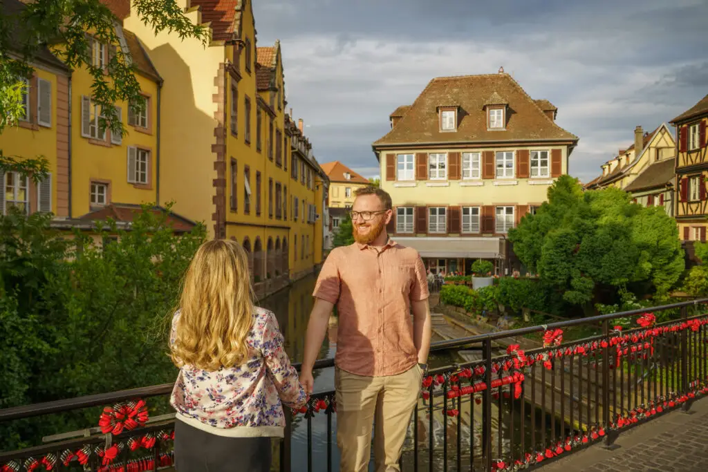 Couple standing on a love lock bridge in La Petite Venise, Colmar, with picturesque views of the Lauch river and traditional Alsatian architecture, perfect for romantic and cultural travel experiences in France.
