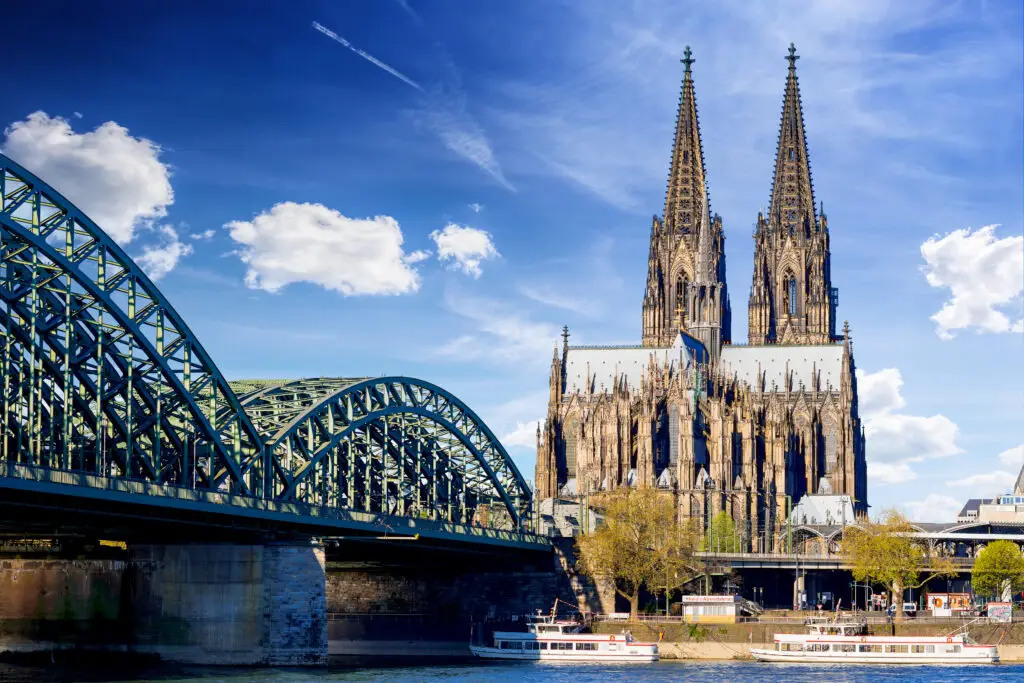 Cologne Cathedral towering next to the Hohenzollern Bridge over the Rhine River in Germany, under a clear blue sky.