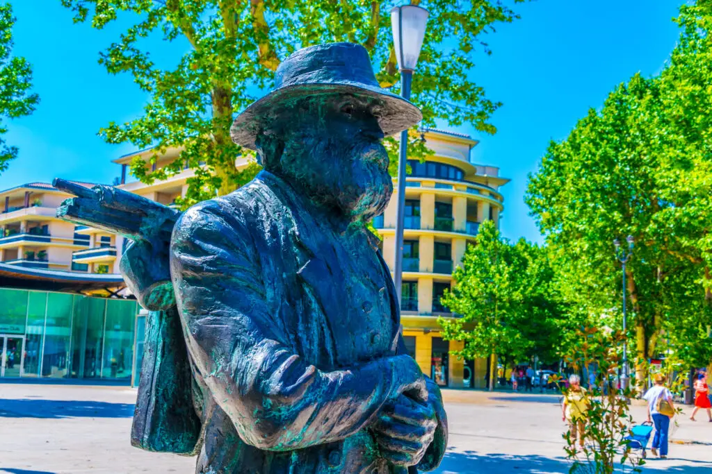 Bronze statue of Paul Cezanne in Aix-en-Provence, France, capturing the artist in mid-pose with a vibrant backdrop of green trees and modern architecture.