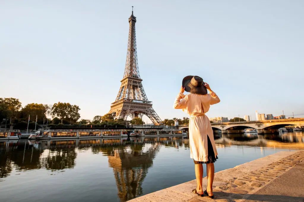 Woman in stylish outfit admiring the Eiffel Tower from a riverside in Paris, highlighting a quintessential tourist experience in France.