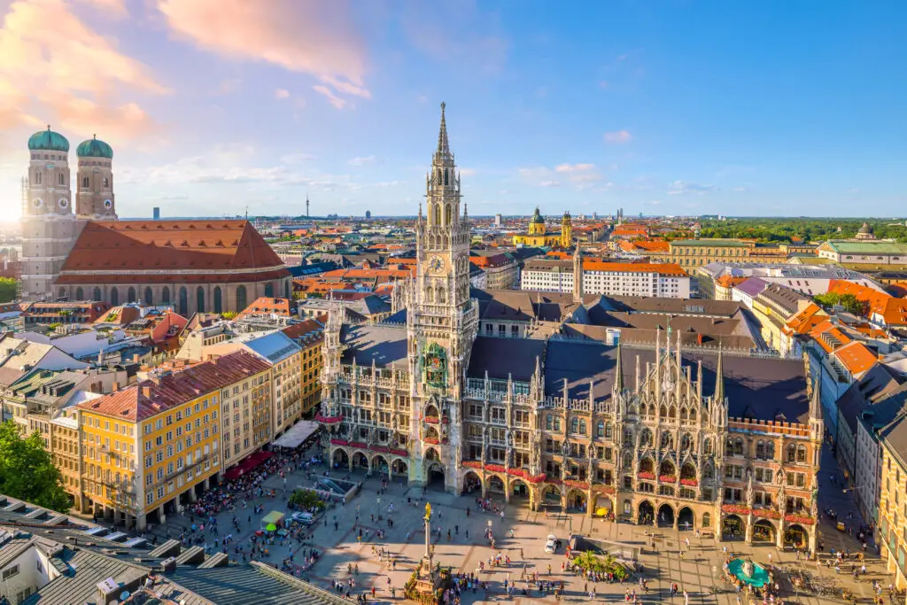 Aerial view of Munich showing the Marienplatz town hall with its intricate Gothic architecture, surrounded by bustling squares and historical buildings under a clear sky.