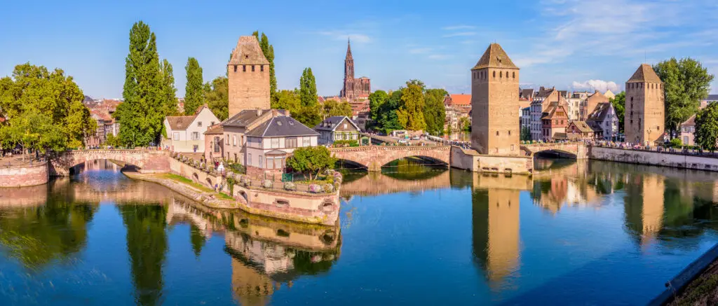 Panoramic view of the historic Ponts Couverts and reflections on the Ill River in Strasbourg, France, showcasing medieval towers and a bridge with a backdrop of traditional Alsatian architecture.