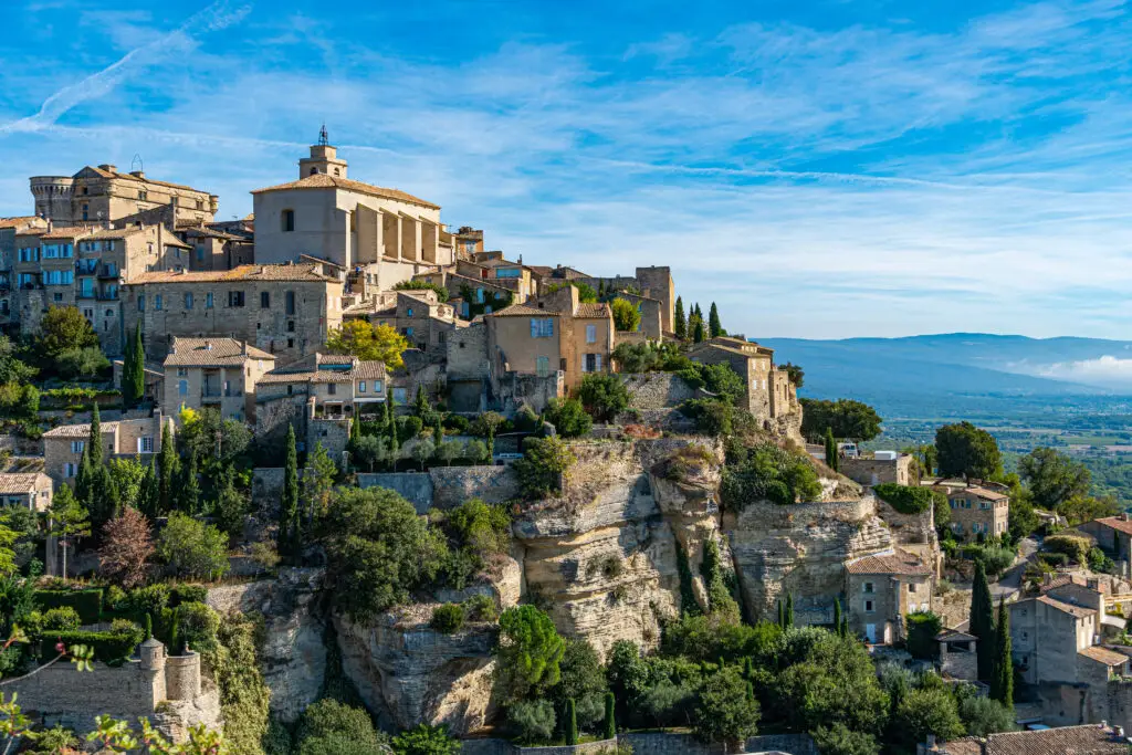 Panoramic view of Gordes, a picturesque hilltop town in Provence, France, showcasing historic stone buildings and lush greenery under a clear blue sky.