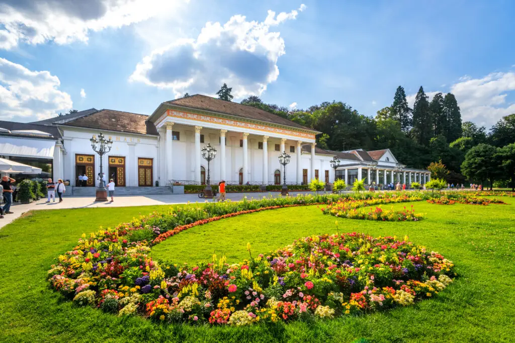 Visitors outside the neoclassical Kurhaus of Baden-Baden, Germany, surrounded by vibrant floral gardens on a sunny day