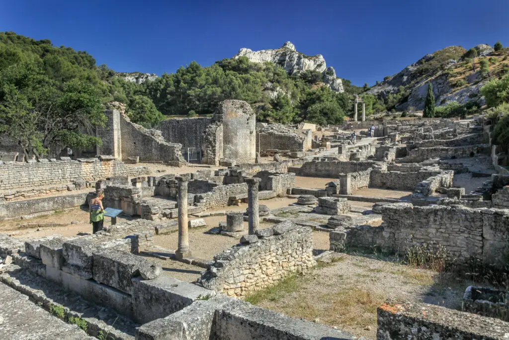 Tourists exploring the ancient Roman ruins of Glanum in Provence, St. Remy, France, surrounded by lush greenery and rugged mountains.