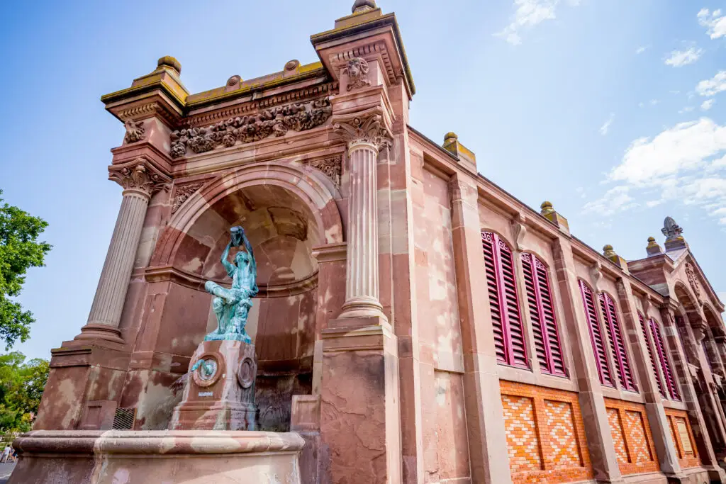 Bronze statue and ornate facade of the covered market in Colmar, France, under a clear blue sky.