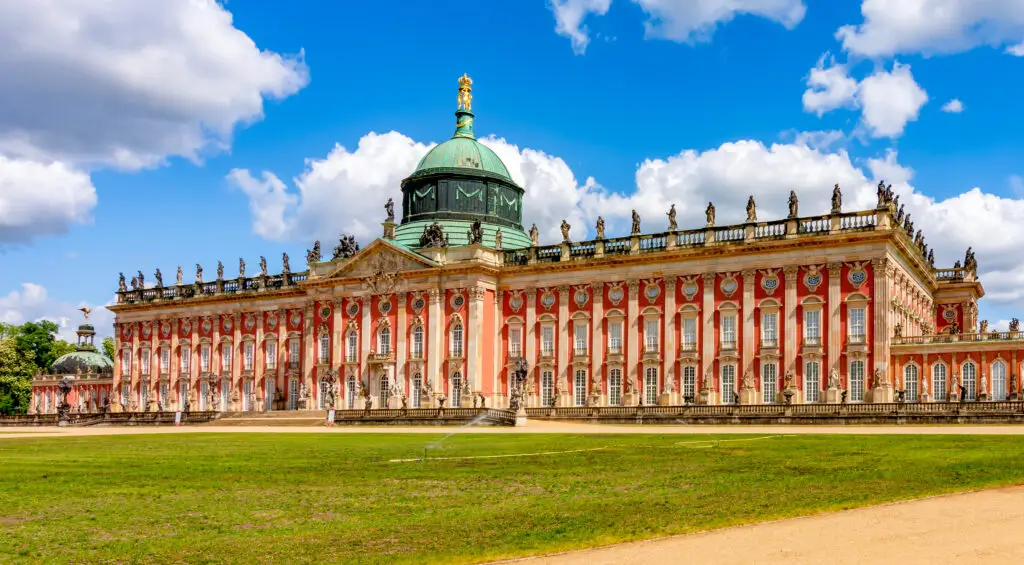 Panoramic view of the ornate New Palace (Neues Palais) with its red and white facade and green dome, set against a bright blue sky in Sanssouci Park, Potsdam, Germany.