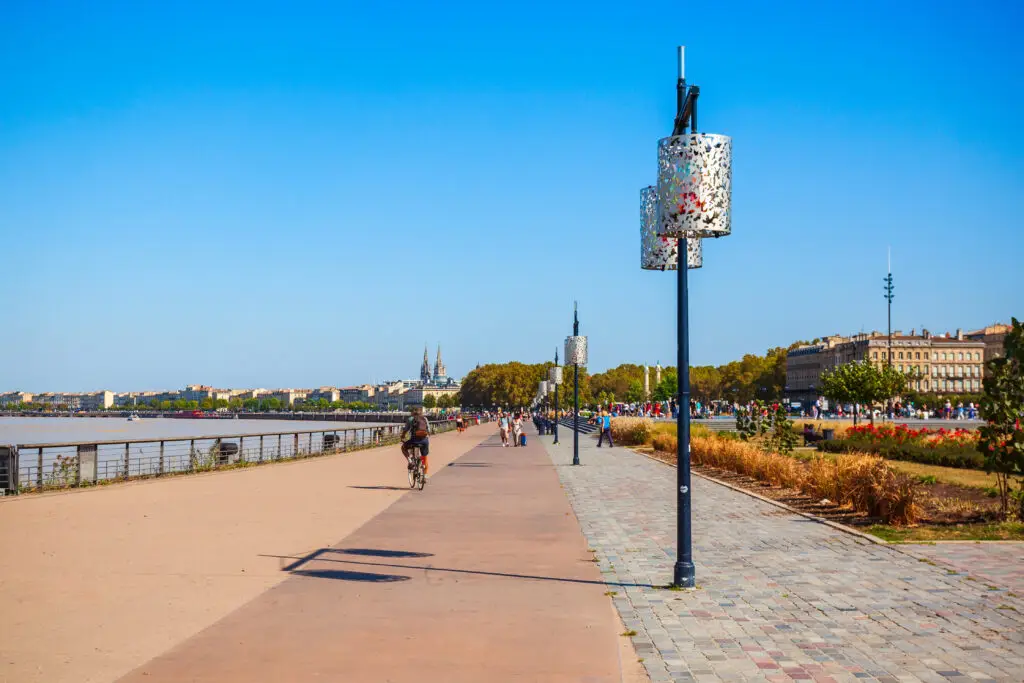 Scenic view of the embankment along the Garonne River in Bordeaux, France, featuring pedestrians and cyclists enjoying a sunny day near historical city buildings.