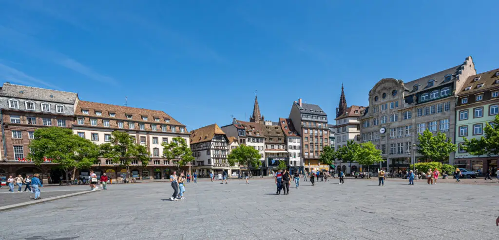 Vibrant scene at Place Kleber, Strasbourg showing tourists and traditional French architecture under blue sky.