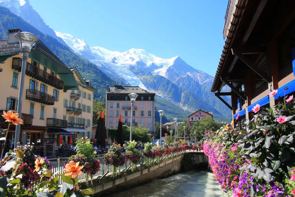 Scenic view of Chamonix Mont Blanc town, showing vibrant flowers along a canal with traditional alpine architecture and snowy mountain backdrop in the French Alps, Haute Savoie.