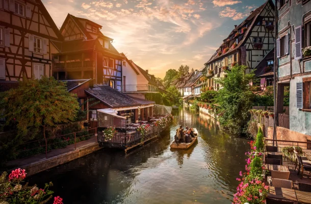 Tourists enjoying a serene boat ride at Fisherman's Wharf in La Petite Venise, Colmar, France, surrounded by traditional half-timbered houses and vibrant flowers at sunset.
