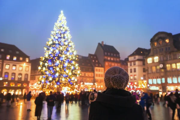 Woman admiring a tall, brightly lit Christmas tree at a bustling market in Strasbourg, France, showcasing festive New Year lights and joyous holiday atmosphere.