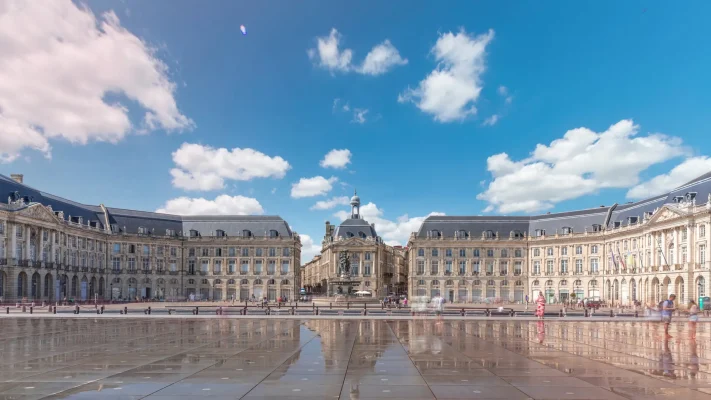 Timelapse view of Place de la Bourse and Miroir d'eau in Bordeaux, France, showcasing the reflective water mirror and historic architecture under a blue sky with clouds.