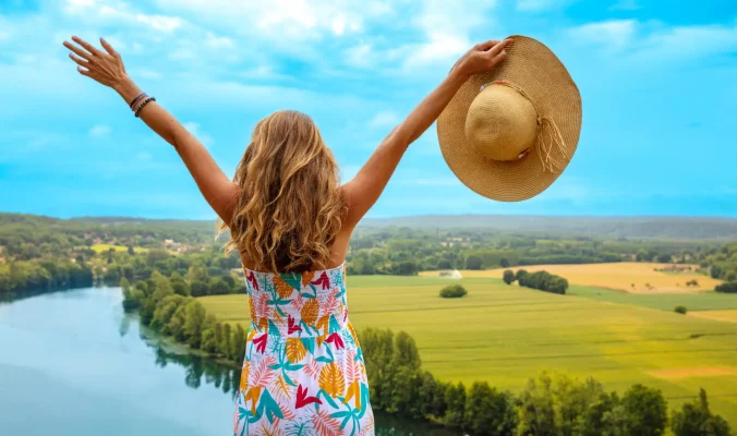 Woman in colorful dress raising arms to embrace panoramic view of Dordogne countryside in France, symbolizing freedom and travel exploration.