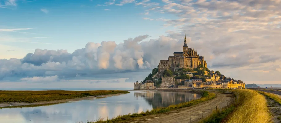 Panoramic view of Mont Saint-Michel at sunrise with reflections on the water and a dramatic sky, highlighting the unique architecture and scenic beauty of this iconic French landmark.